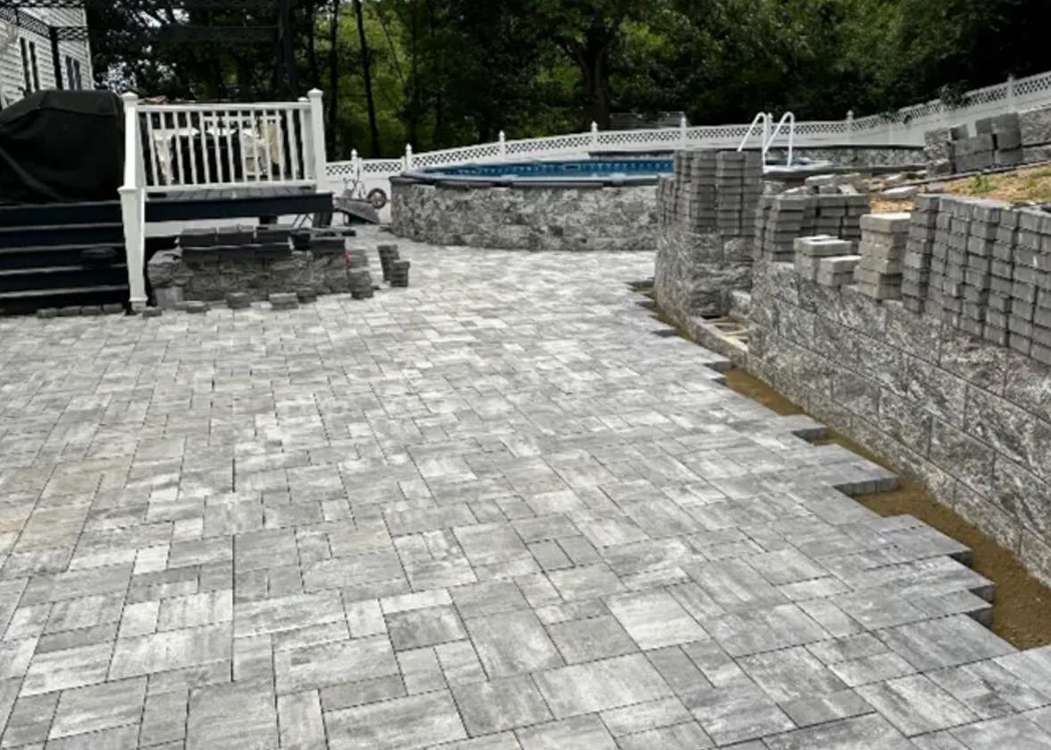Stone patio under construction with a pool and deck. Gray bricks are laid with stacked stone retaining walls.