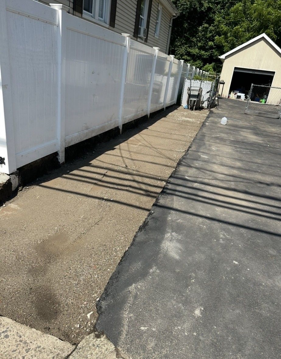 View of a gravel and asphalt surface along a white fence. A garage is in the background.