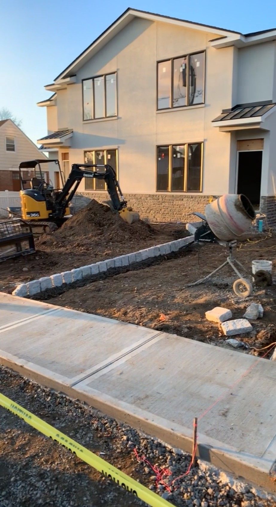 Construction site: excavator next to a building. Concrete sidewalk and curbing in progress, cement mixer visible.