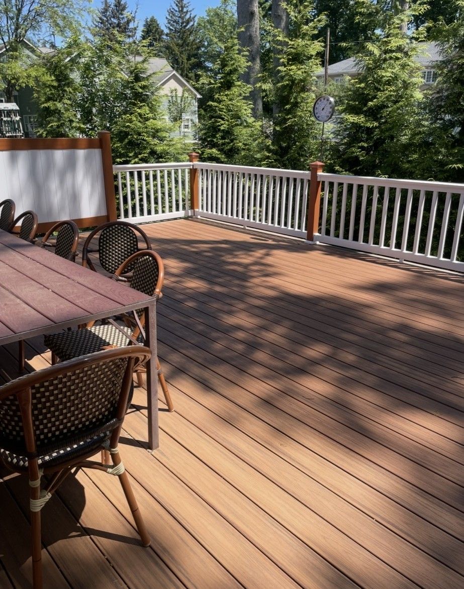 Wooden deck with table and chairs, white railing, and surrounding trees in a sunny outdoor setting.