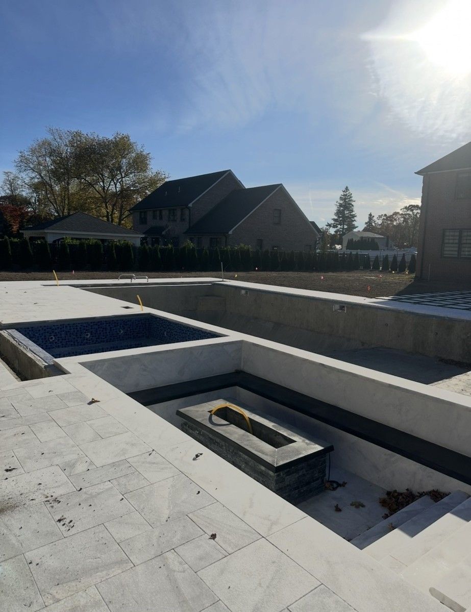 Unfinished backyard pool with fire pit, steps, and surrounding patio; blue sky with sun.