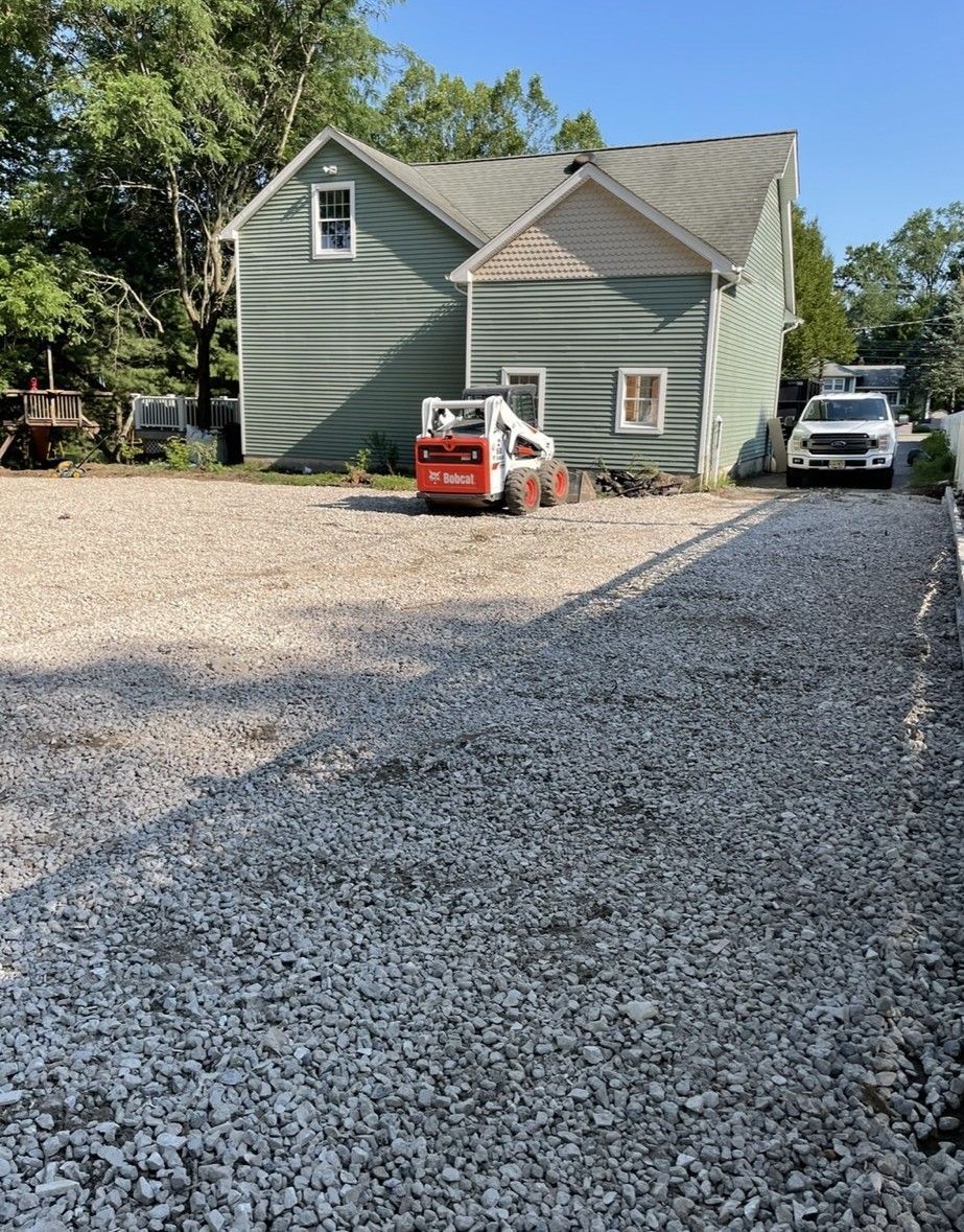 Gravel driveway with a teal house, a skid steer, and a white truck parked. Trees are in the background.