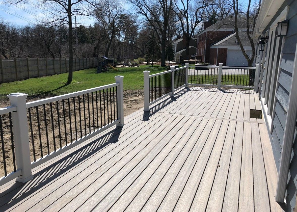 Wooden deck with white railing and black spindles, overlooking a grassy yard with trees and a house.