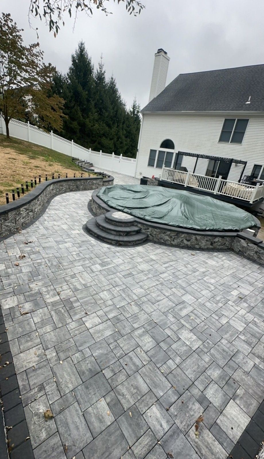 Gray and black paver patio with a covered pool and stone retaining walls. House in the background.