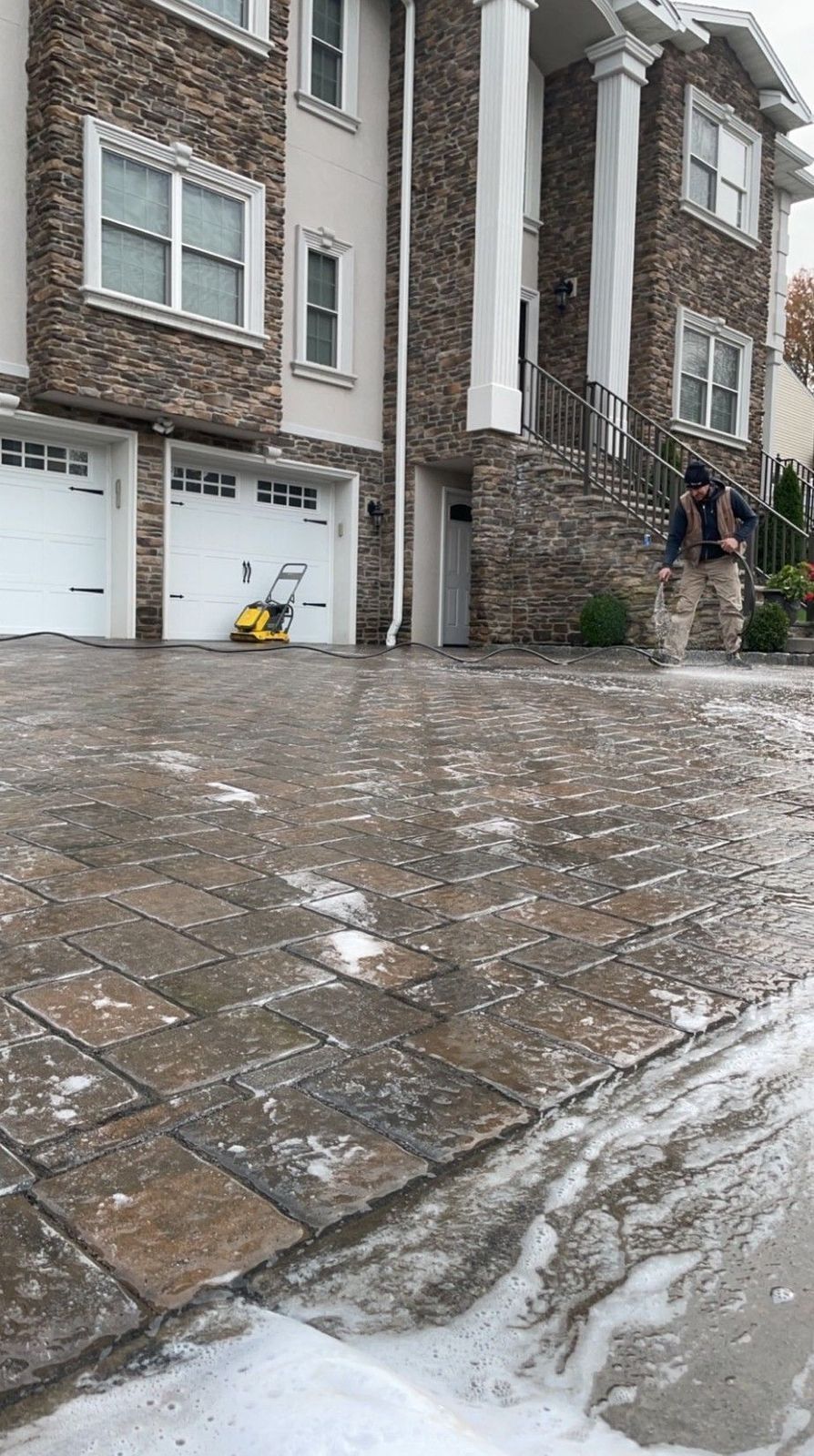 Man power washing a brick driveway in front of a multi-story house.