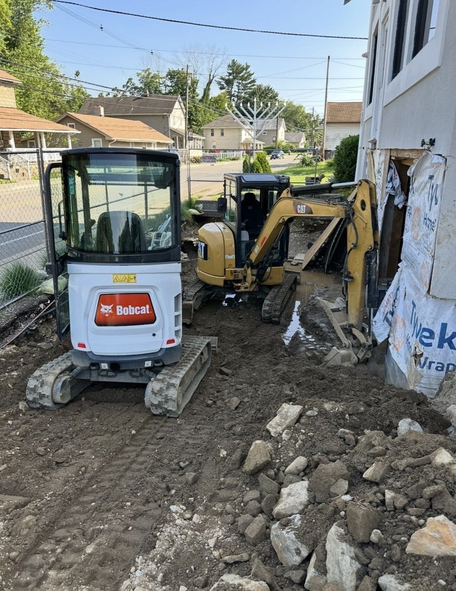 Two excavators next to a building, digging in the dirt. Sunlight.