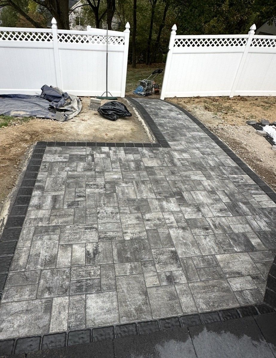 Brick paver patio and walkway with white fence in the background. Construction materials and tools are present.