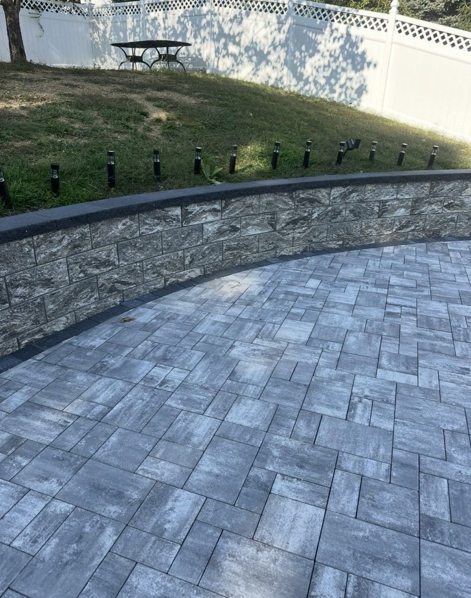 Curved gray brick patio and retaining wall, grassy hill with lights, bench, white wall in background.