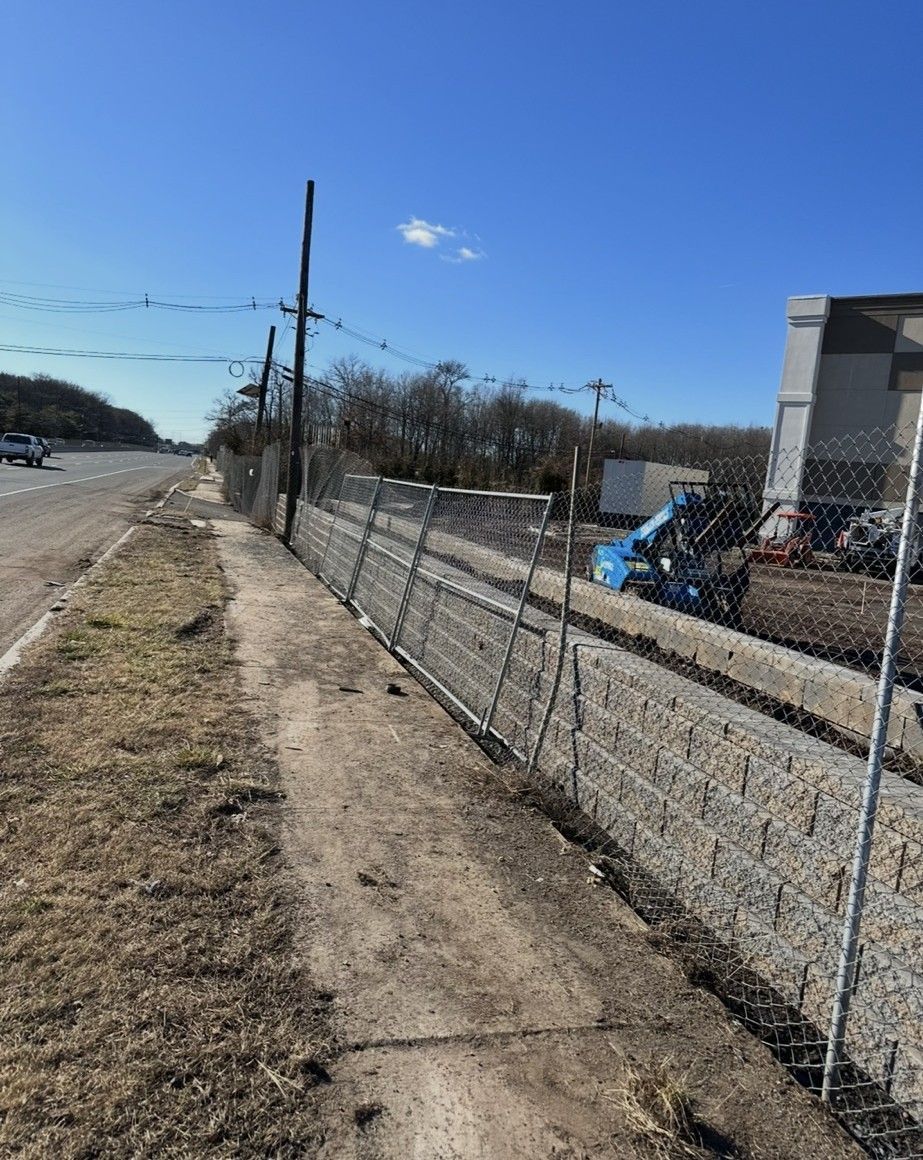 Sidewalk next to highway, chain-link fence, utility poles, construction site visible. Bright blue sky.
