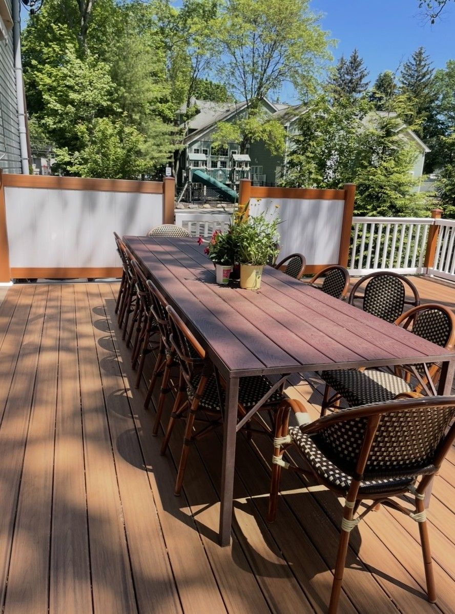 Wooden deck with long table and chairs; greenery and homes in the background. Sunny day.