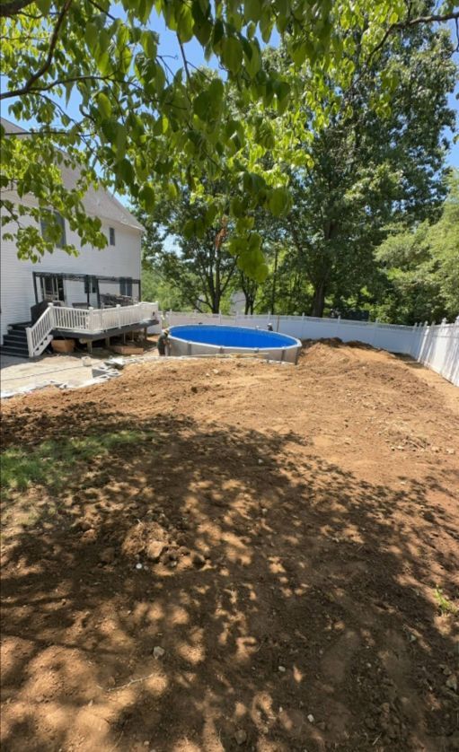 Backyard with above-ground pool, dirt mound, white fence, and house. Blue water in pool. Sunny day.