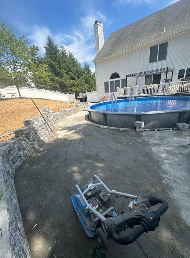 Backyard with above-ground pool and construction equipment; dirt ground being compacted, retaining wall, house in background.