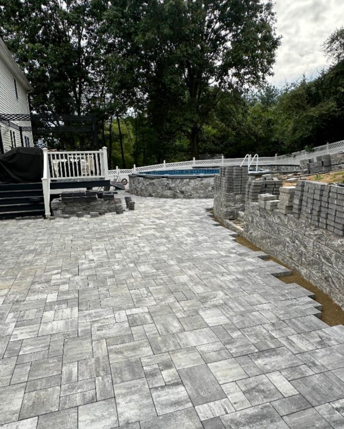 Stone patio being constructed around an in-ground pool, next to a deck and green trees.