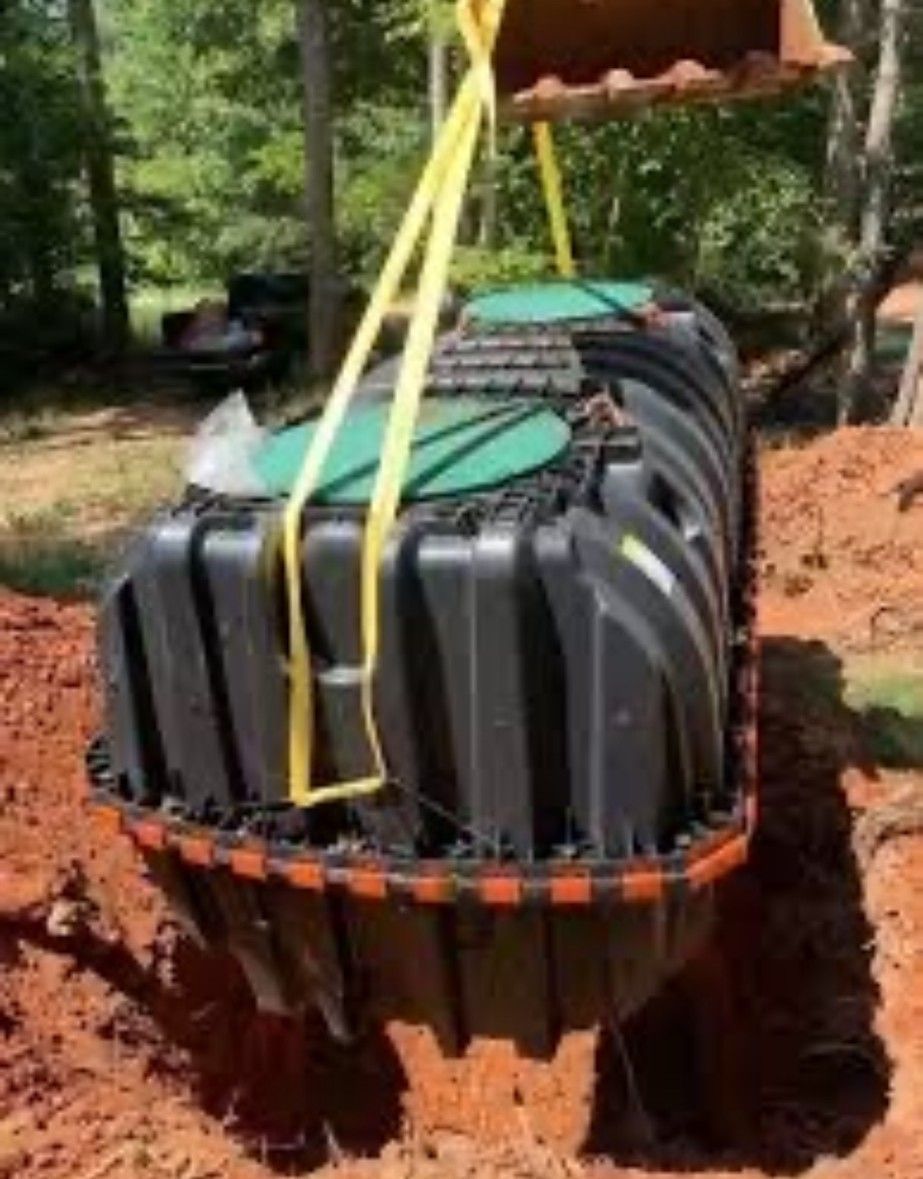 Black septic tank being lowered into an excavated hole by yellow straps attached to a piece of machinery.