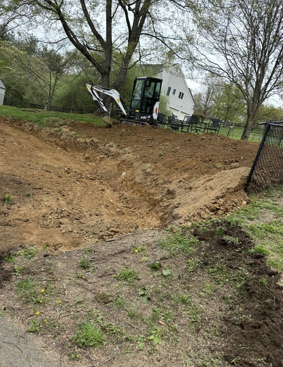 Dirt work in progress; Bobcat compact track loader on a muddy hillside near a house and trees.