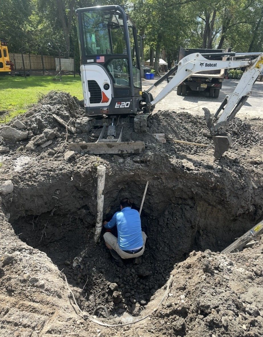 Person in a hole, working near an excavator. Brown dirt surrounds the hole in an outdoor setting.