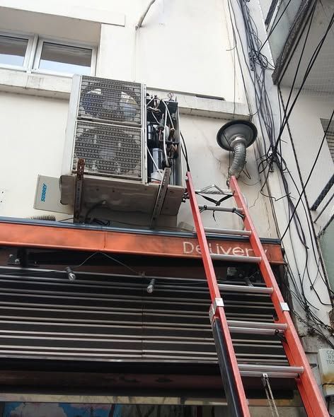 Unidad de aire acondicionado exterior en la pared del edificio con escalera de acceso. Cables eléctricos.