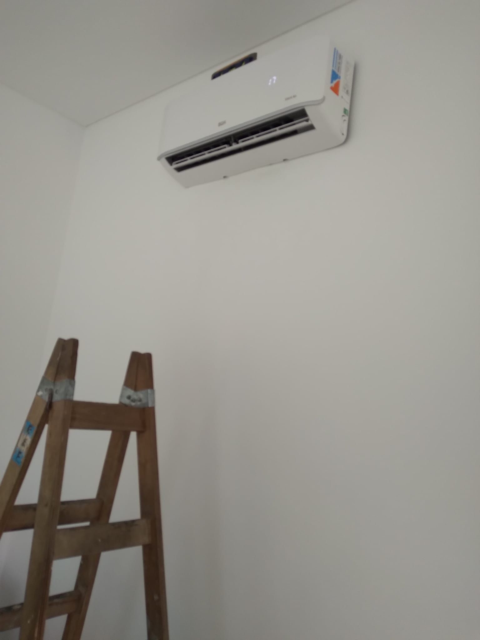 Aire acondicionado montado en pared blanca, escalera de madera en la esquina.