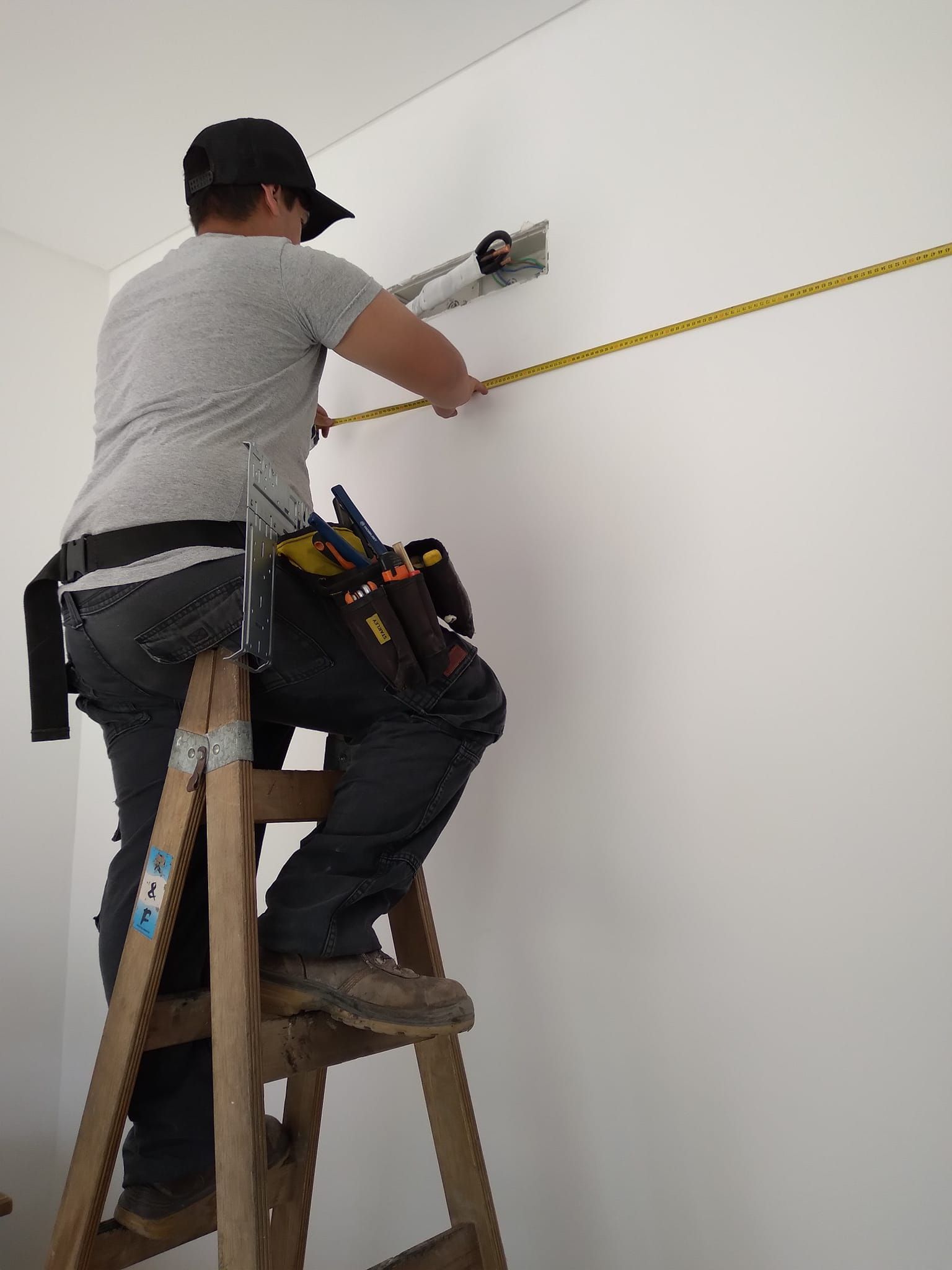 Persona en una escalera midiendo una pared blanca con una cinta métrica amarilla, trabajando en un accesorio de metal.