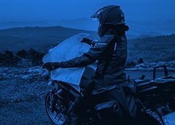 Motociclista com capa de chuva ao lado de uma moto em uma estrada de montanha enevoada ao entardecer.
