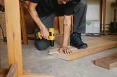 Person kneeling, using a yellow drill on wood in a workshop.
