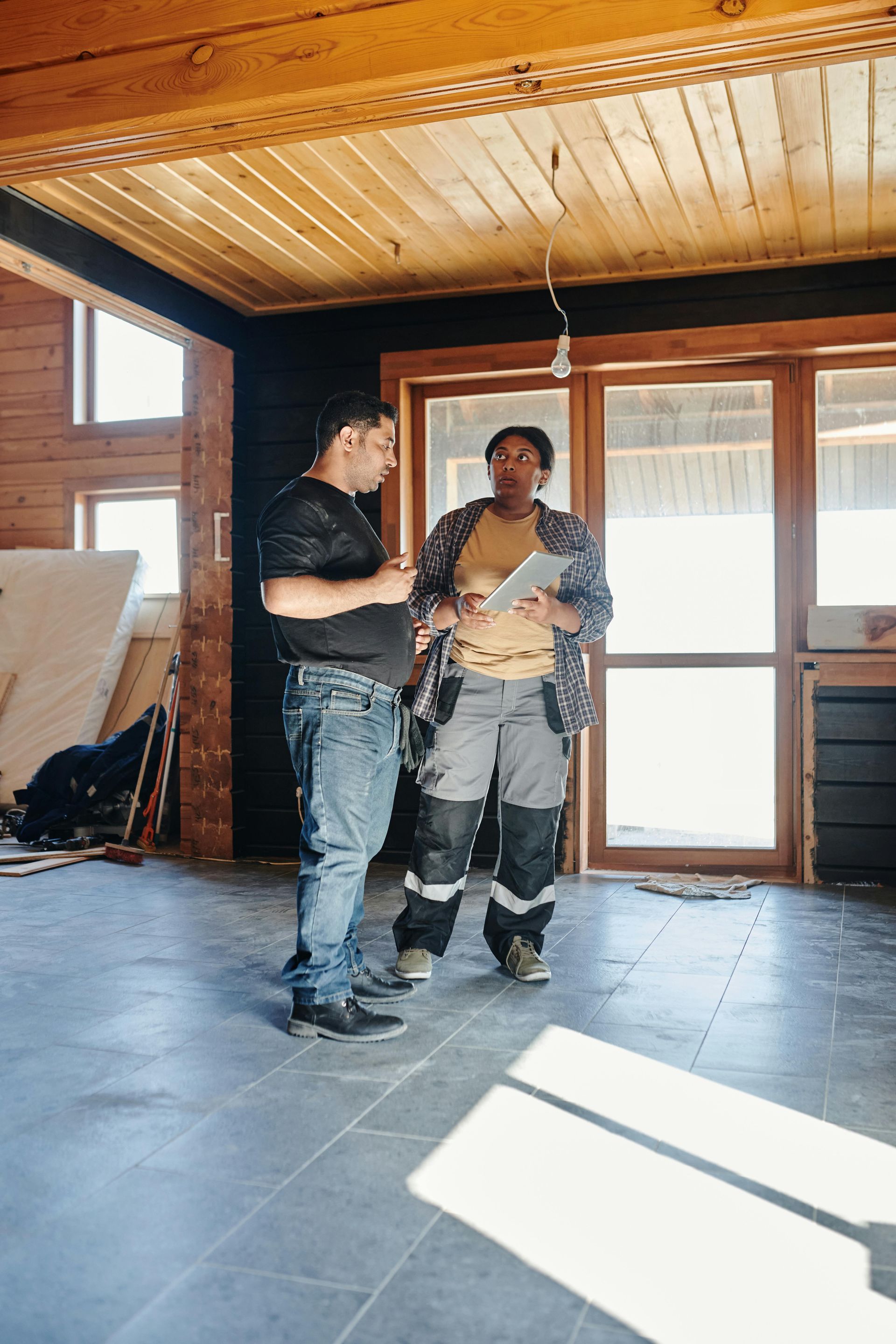 Two people in a room under construction, discussing plans, one holding a tablet, wearing work clothes.