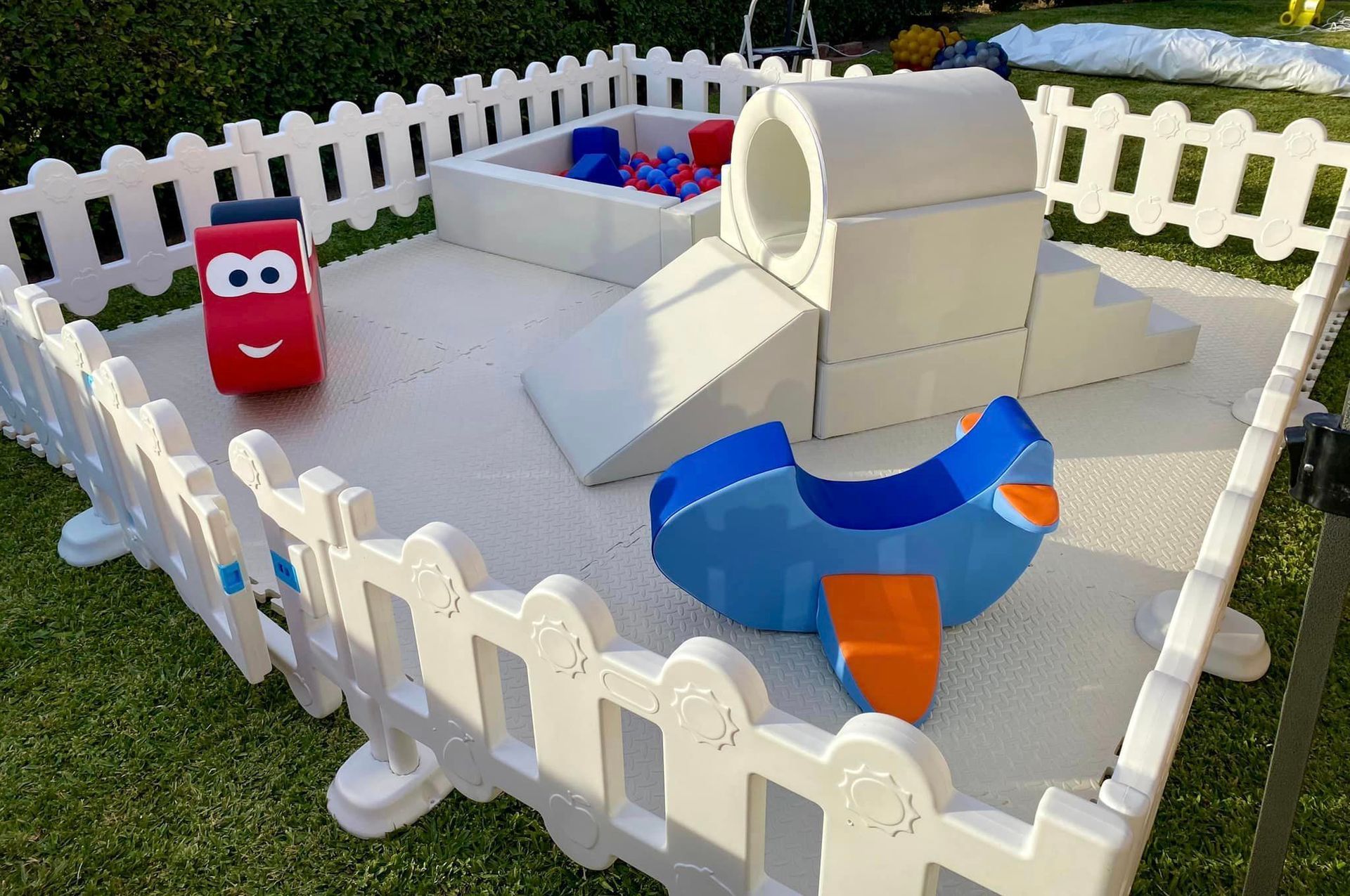 A white fence surrounds a playground with toys and a ball pit.