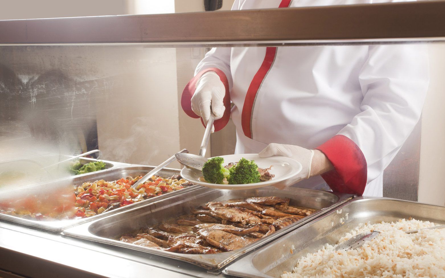 Buffet server plating salad at a stainless-steel food counter with hot dishes and steam
