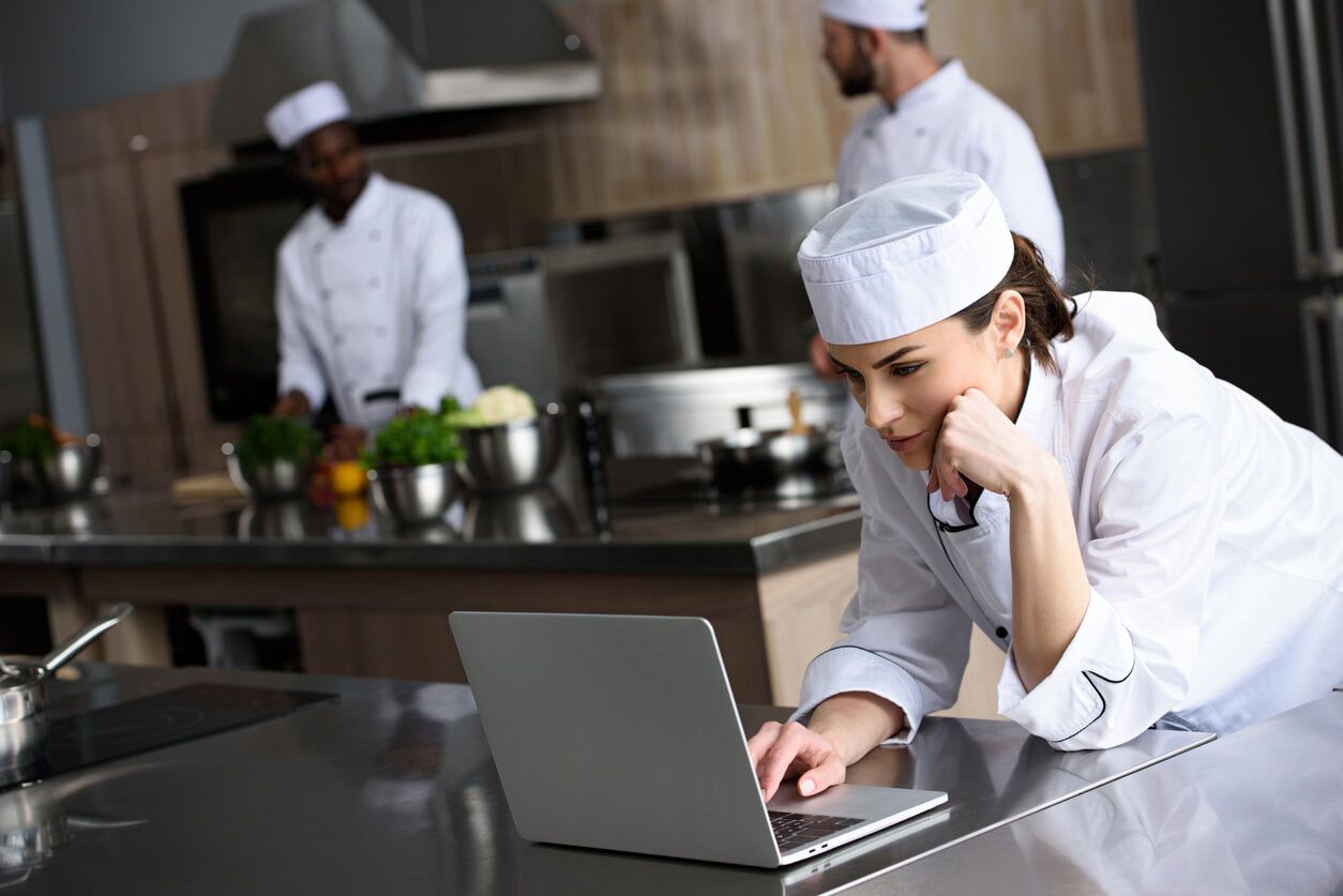 Chef leaning on counter, looking at laptop; other chefs in kitchen.