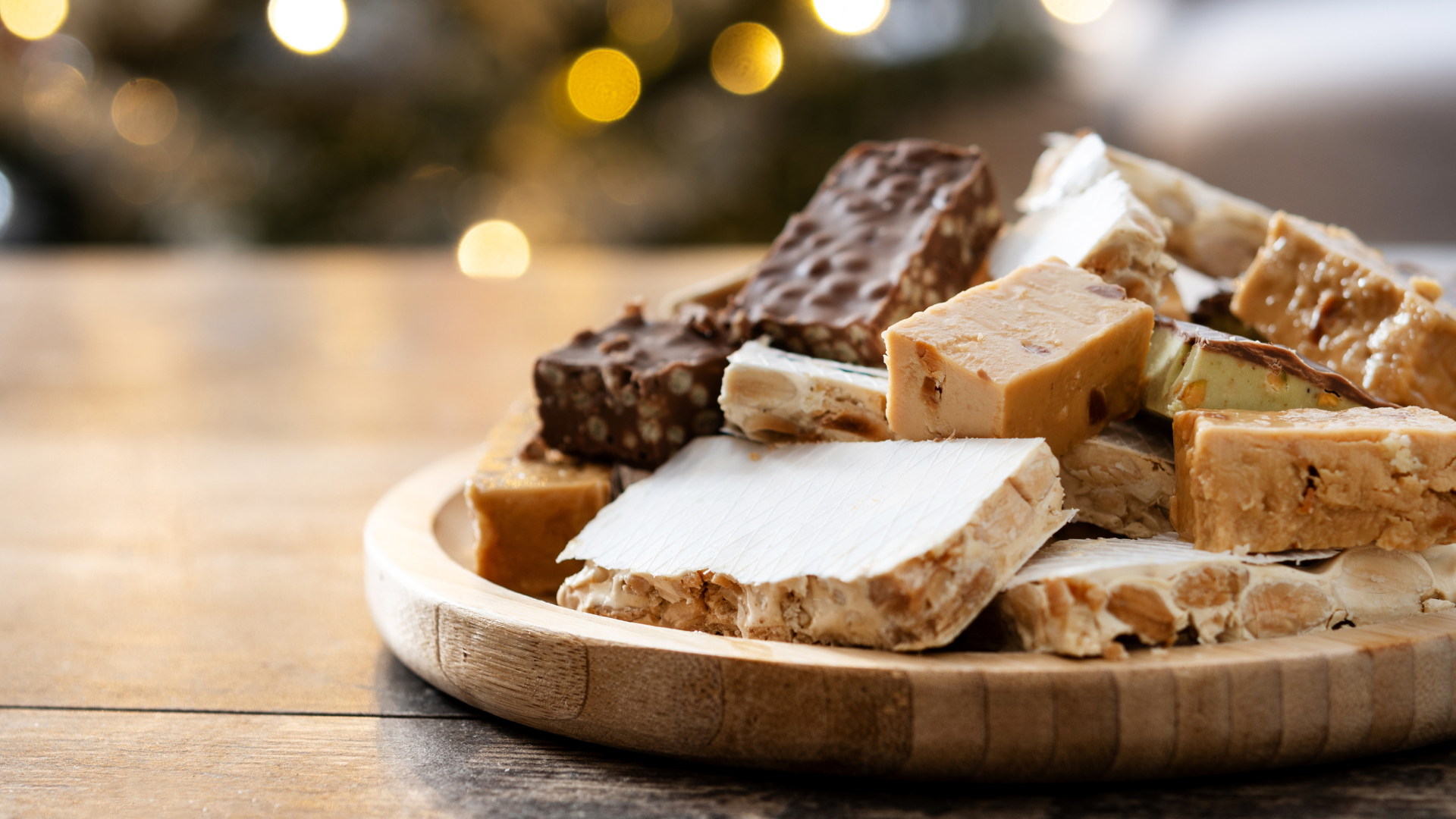 Variety of nougat pieces on a wooden plate, with blurred festive lights in the background.