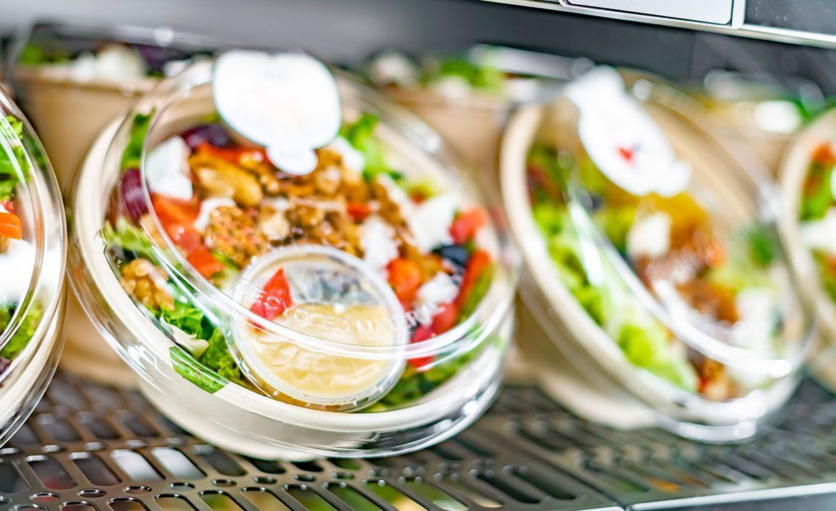 Una fila de ensaladas en recipientes de plástico en un estante de un refrigerador.