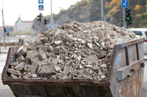 A large, rusted metal dumpster overflowing with gray concrete rubble, set against a blurred city street background.