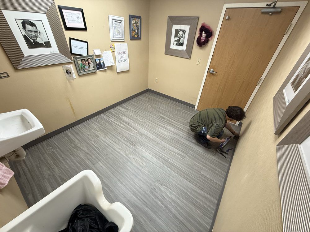 Person crouching by a door in a small clinic room with framed pictures and a sink.