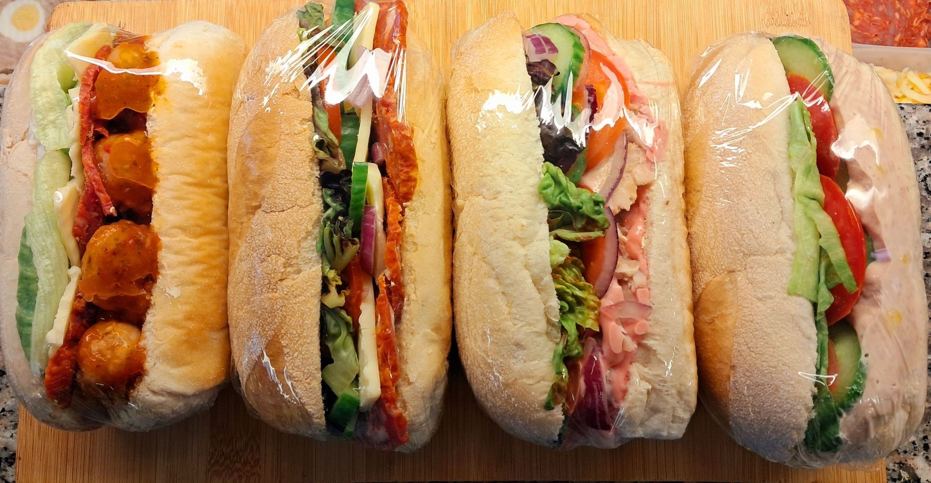 Five wrapped sandwiches with assorted fillings arranged side by side on a wooden surface.