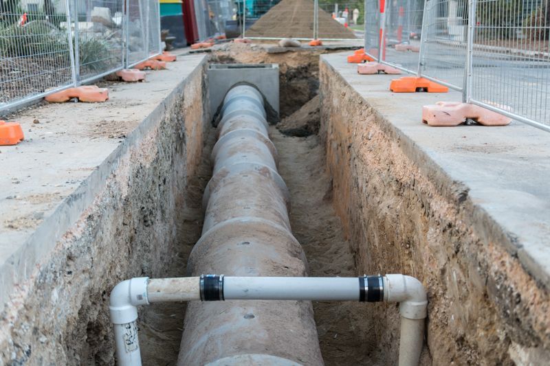 Trench with large concrete pipe, white PVC pipes in foreground, construction site with barriers.
