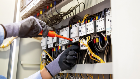 Electrician working on electrical panel, wearing gloves, using a screwdriver, with wires and components visible.
