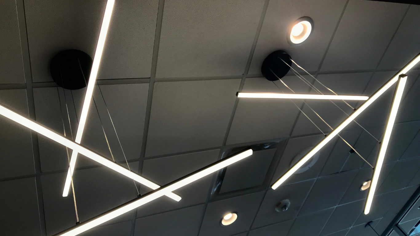 Modern linear pendant lights hang from a grid ceiling. Bright white light against a neutral background.
