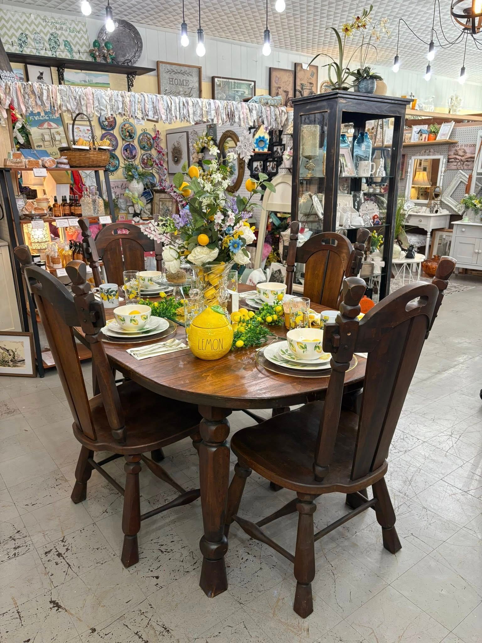 Round wooden dining table with chairs, yellow dishes, and floral decor in a store showroom