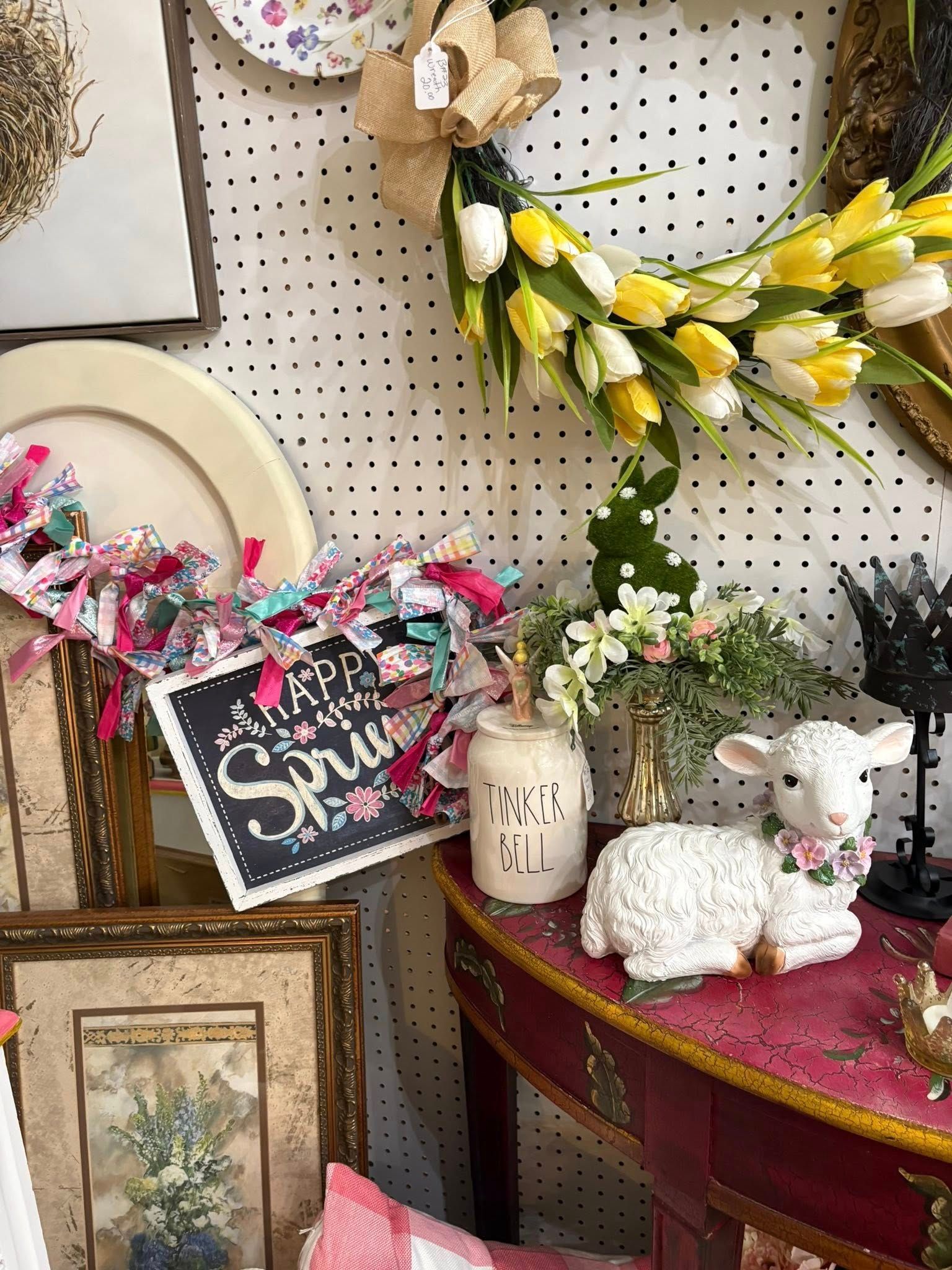 Decorated craft display with floral wreaths, framed art, stuffed bunnies, and a chalkboard sign on a red table