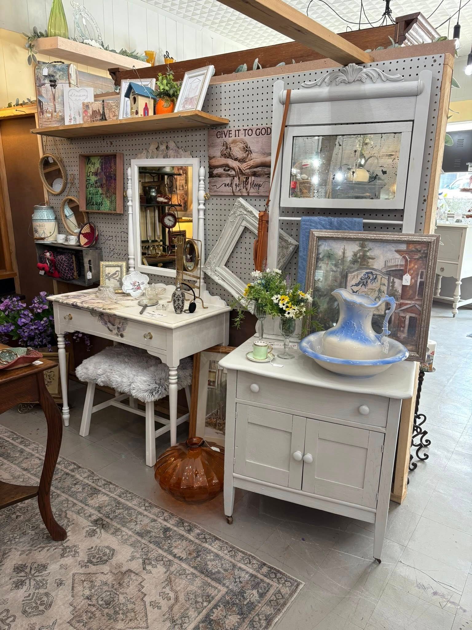 Vintage décor shop display with white vanity, mirror, flowers, lamp, and a small cabinet on a rug-covered floor