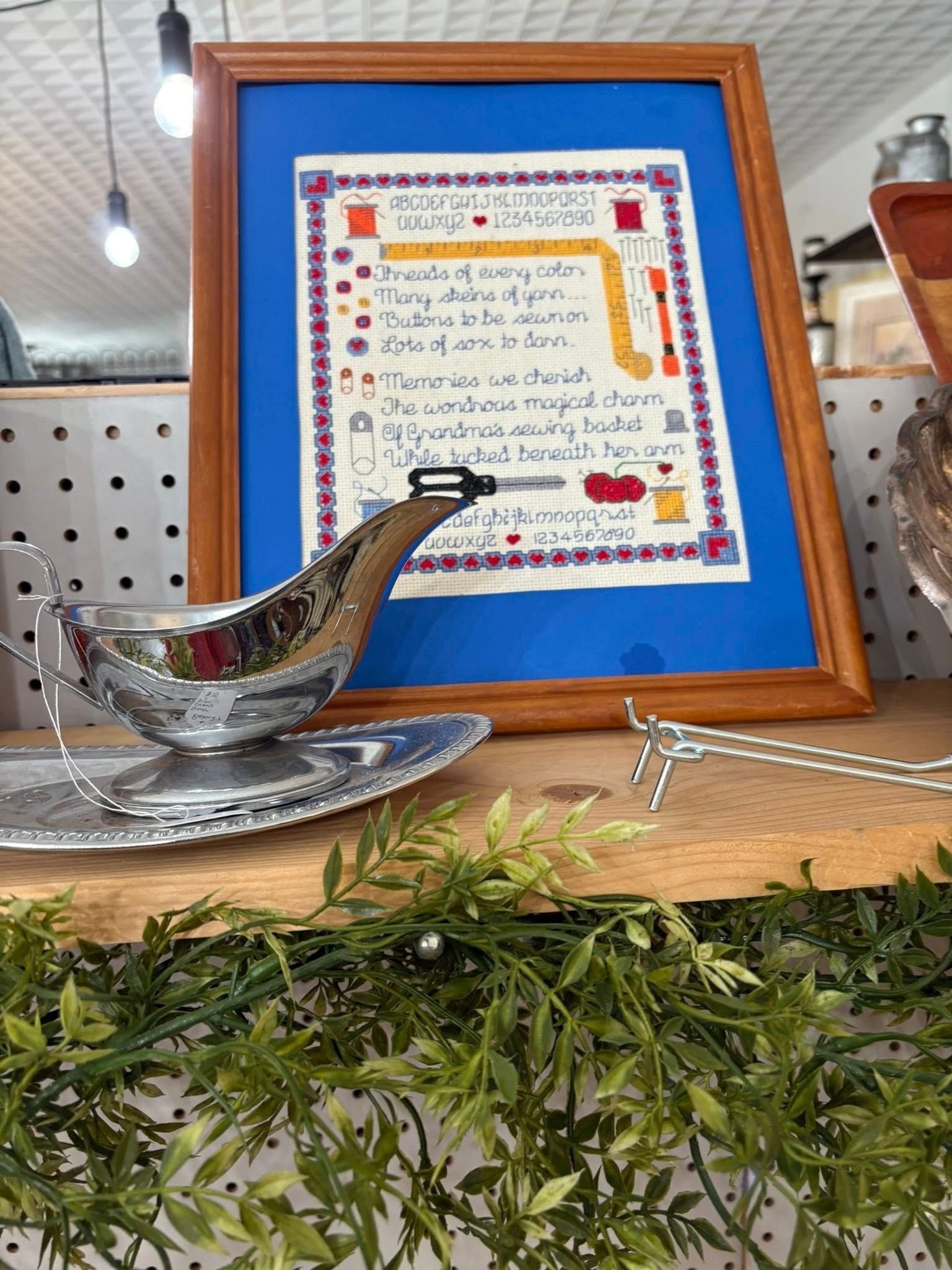 Framed handwritten menu beside a silver gravy boat on a wooden shelf, with greenery in front