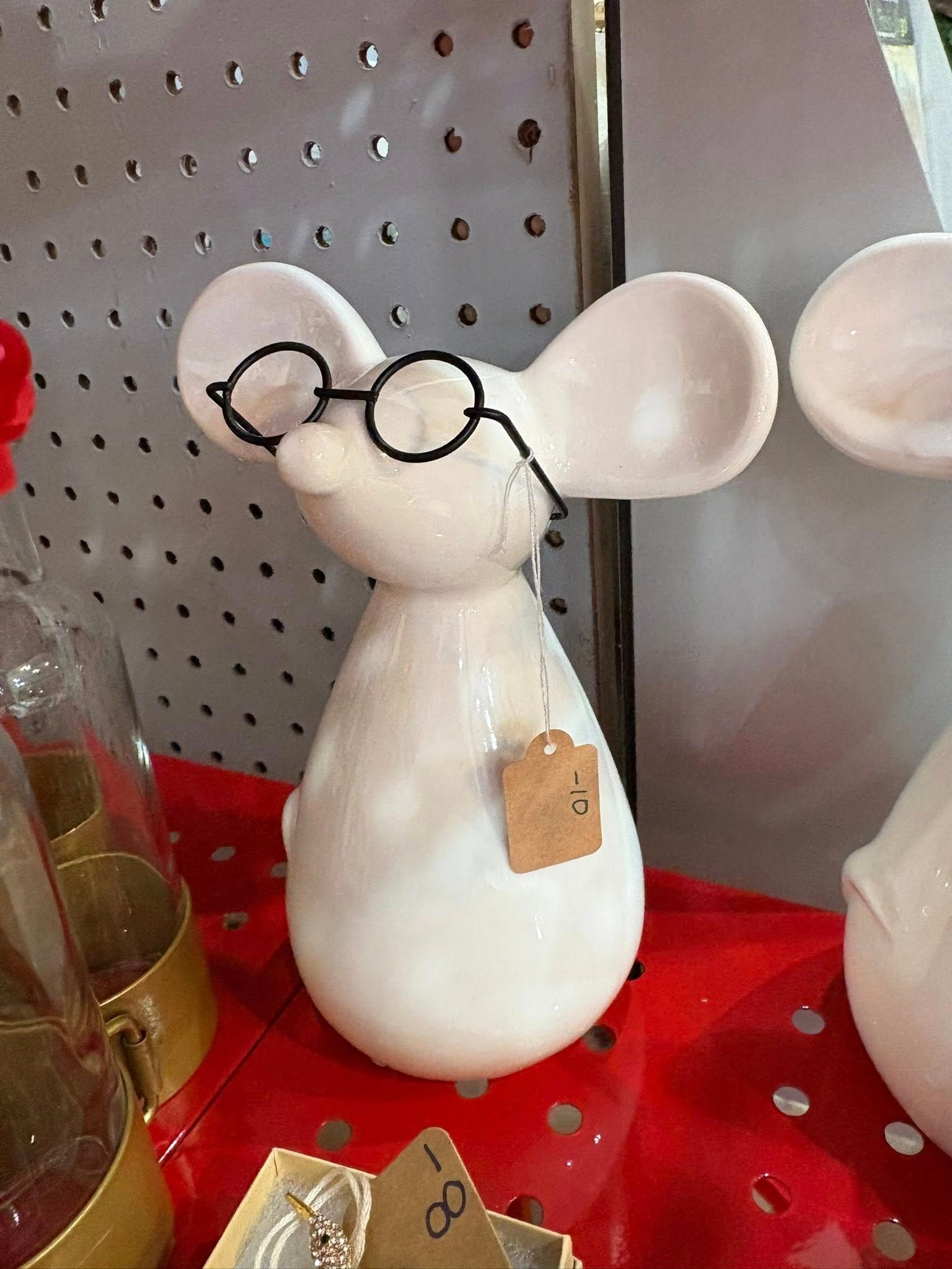 White mouse figurine with round glasses on a red display surface against a gray pegboard background