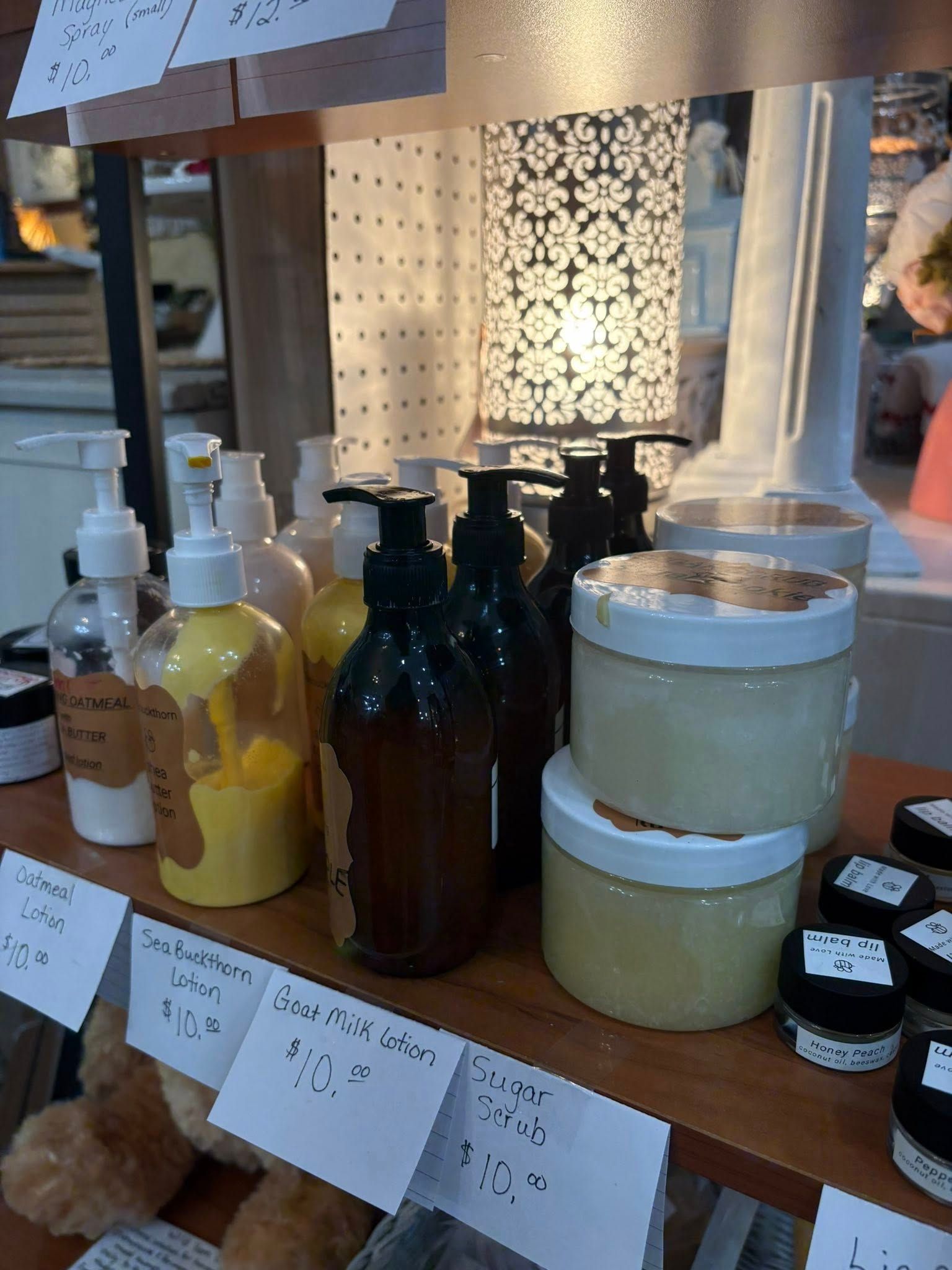 Display of assorted bottled soaps and creams on wooden shelves at a market stall, with price tags and a lamp.