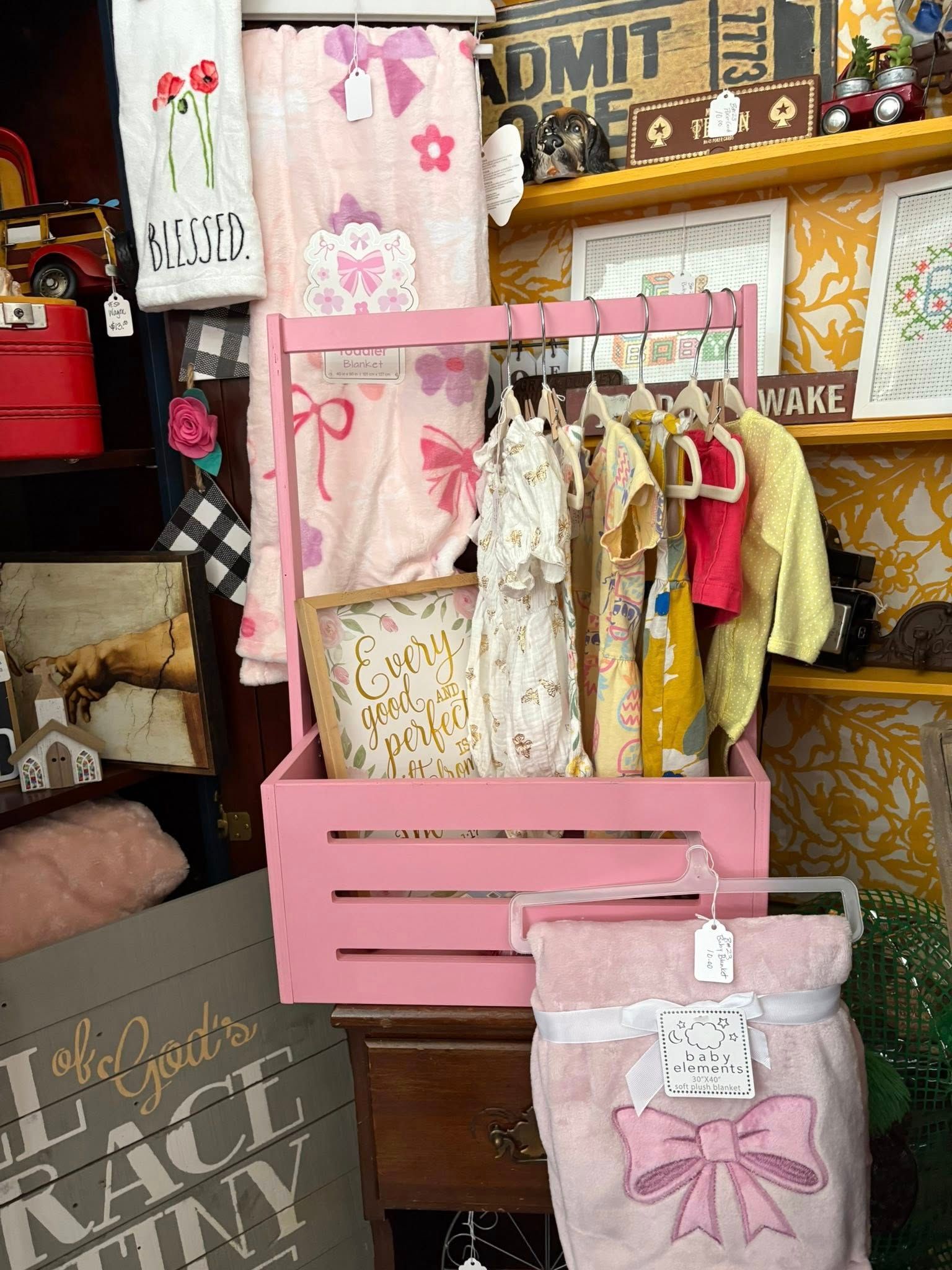 Pink display rack with hanging baby clothes, blankets, and gift items in a store or craft booth