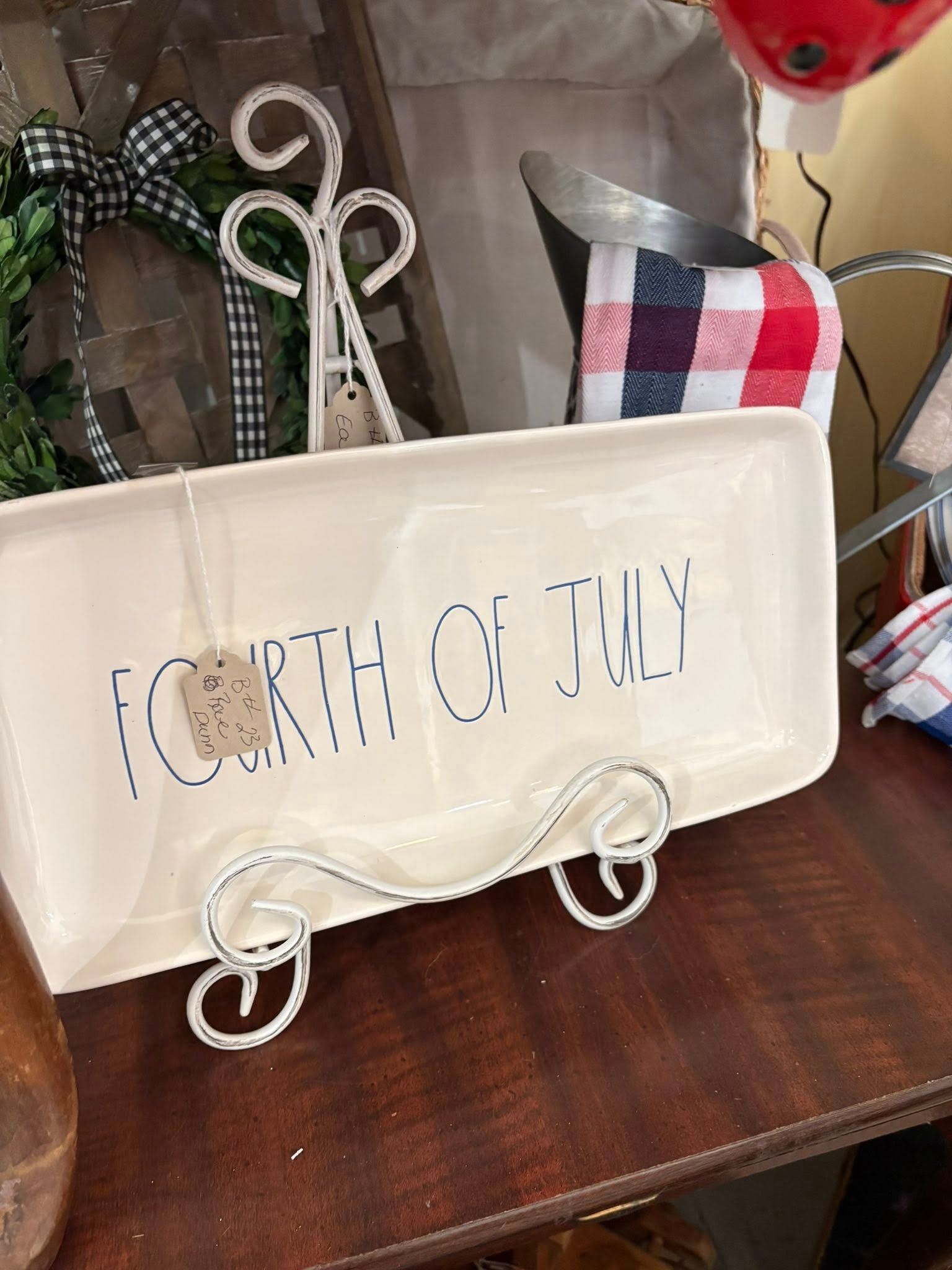 White decorative tray with “Fourth of July” lettering, red-and-blue plaid accents, on a wooden shelf.