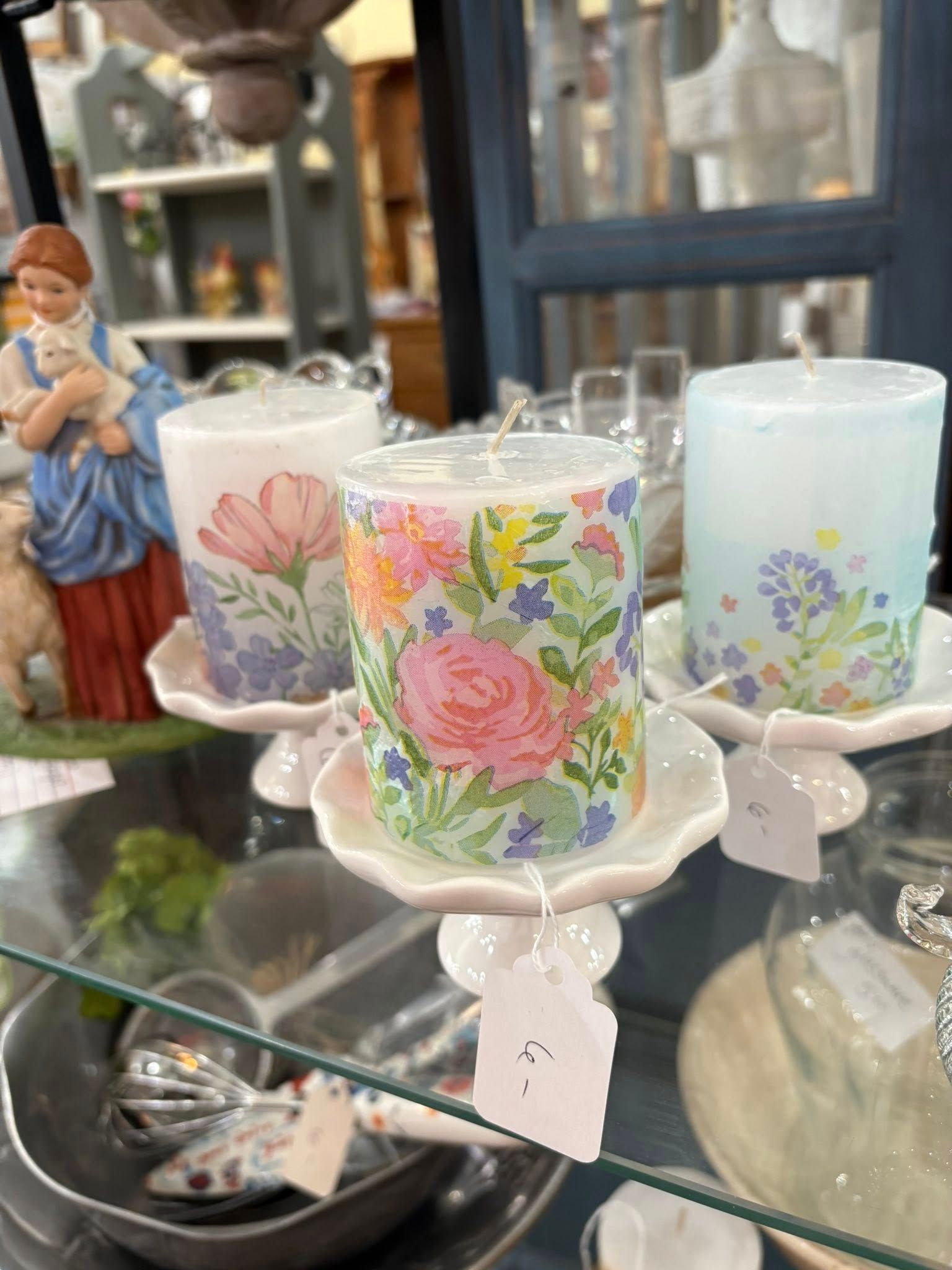 Decorative floral candles in white holders on a glass table, with a small figurine in the background