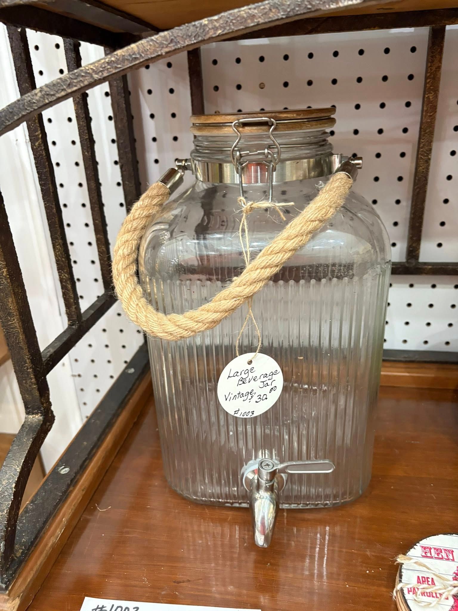 Glass beverage dispenser with rope handle and spigot on a wooden shelf, beside a pegboard wall