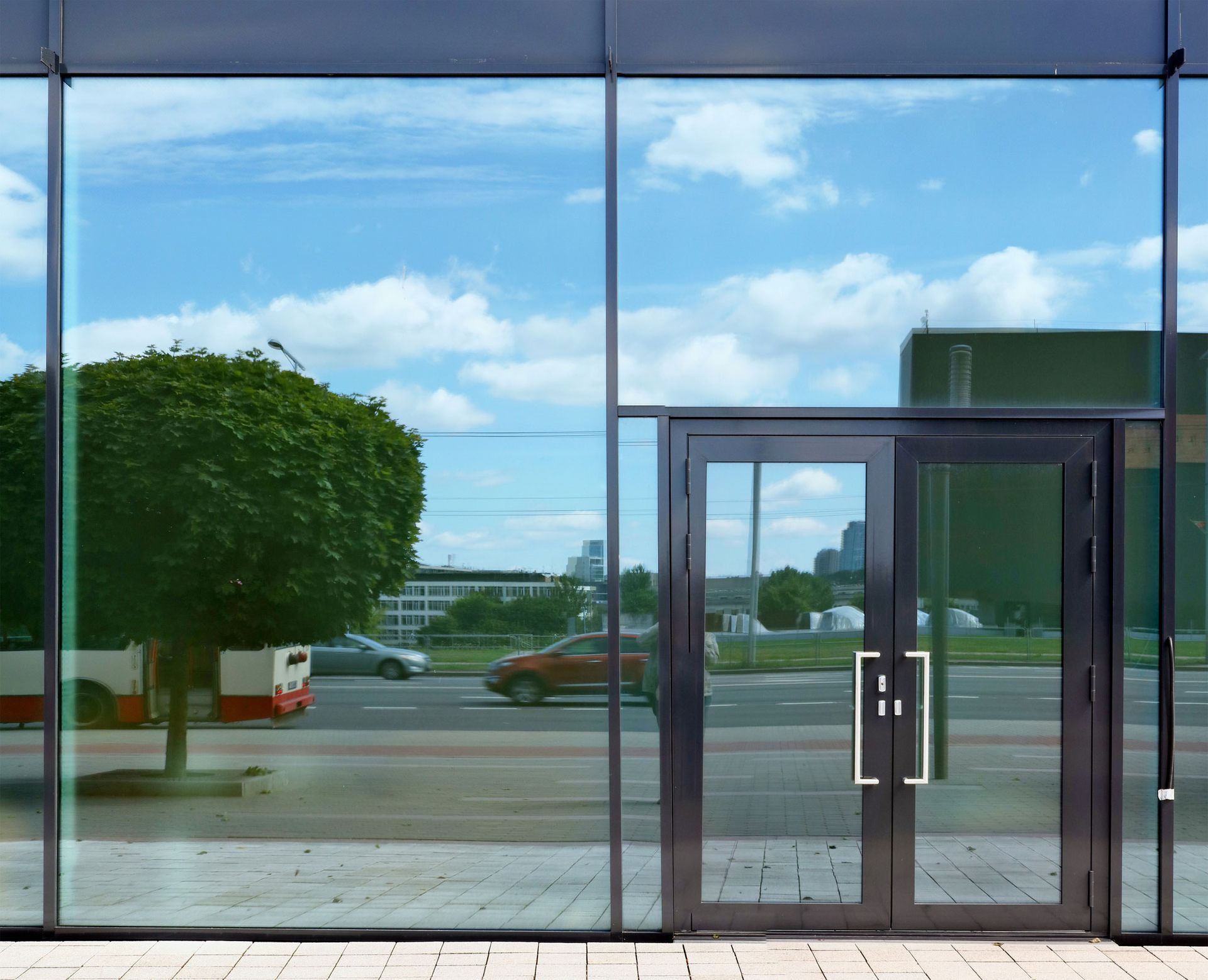 Glass building facade with reflecting sky, street, and vehicles, beside a double door.