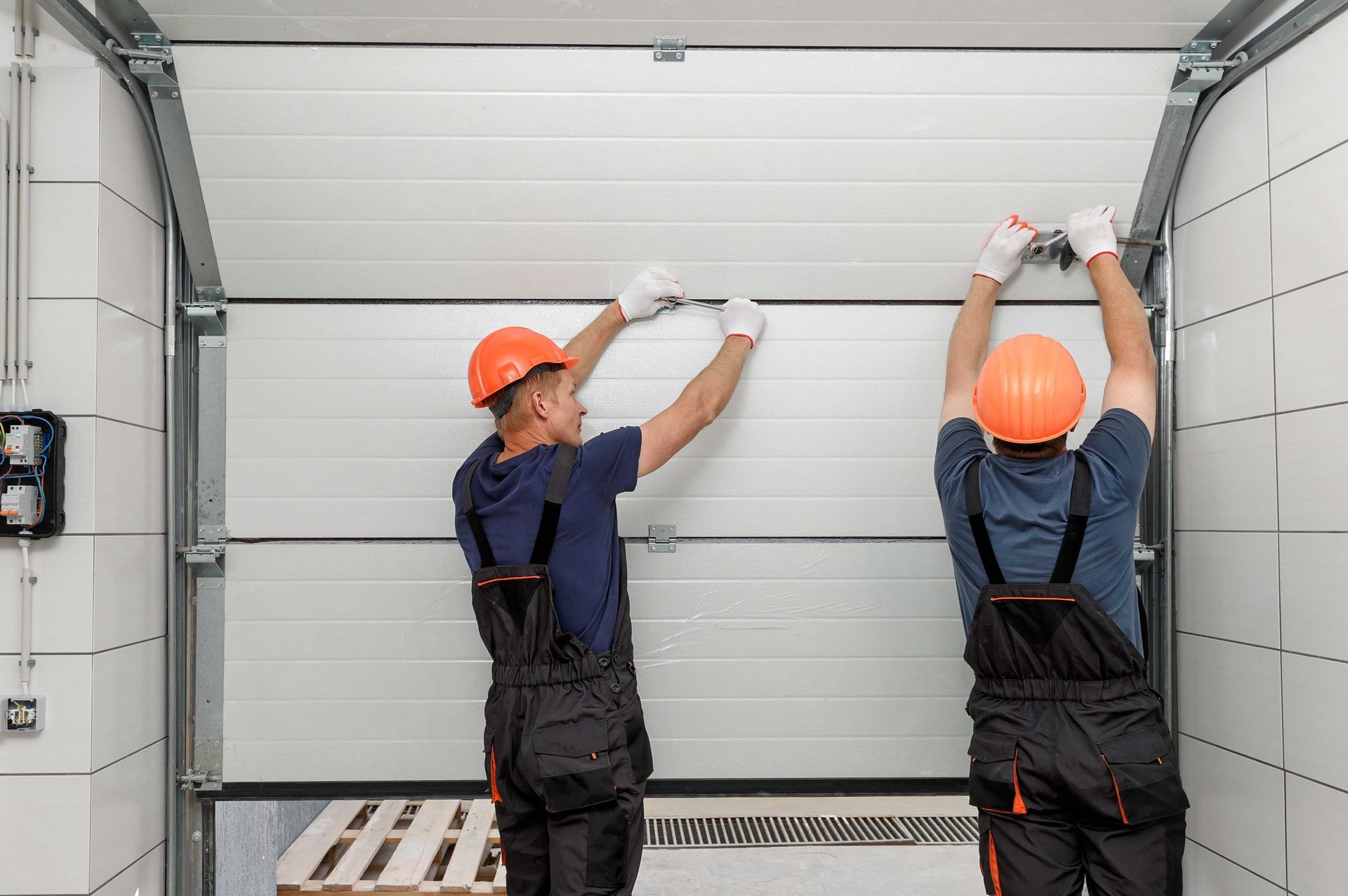 Two workers in overalls and hard hats installing a white garage door.