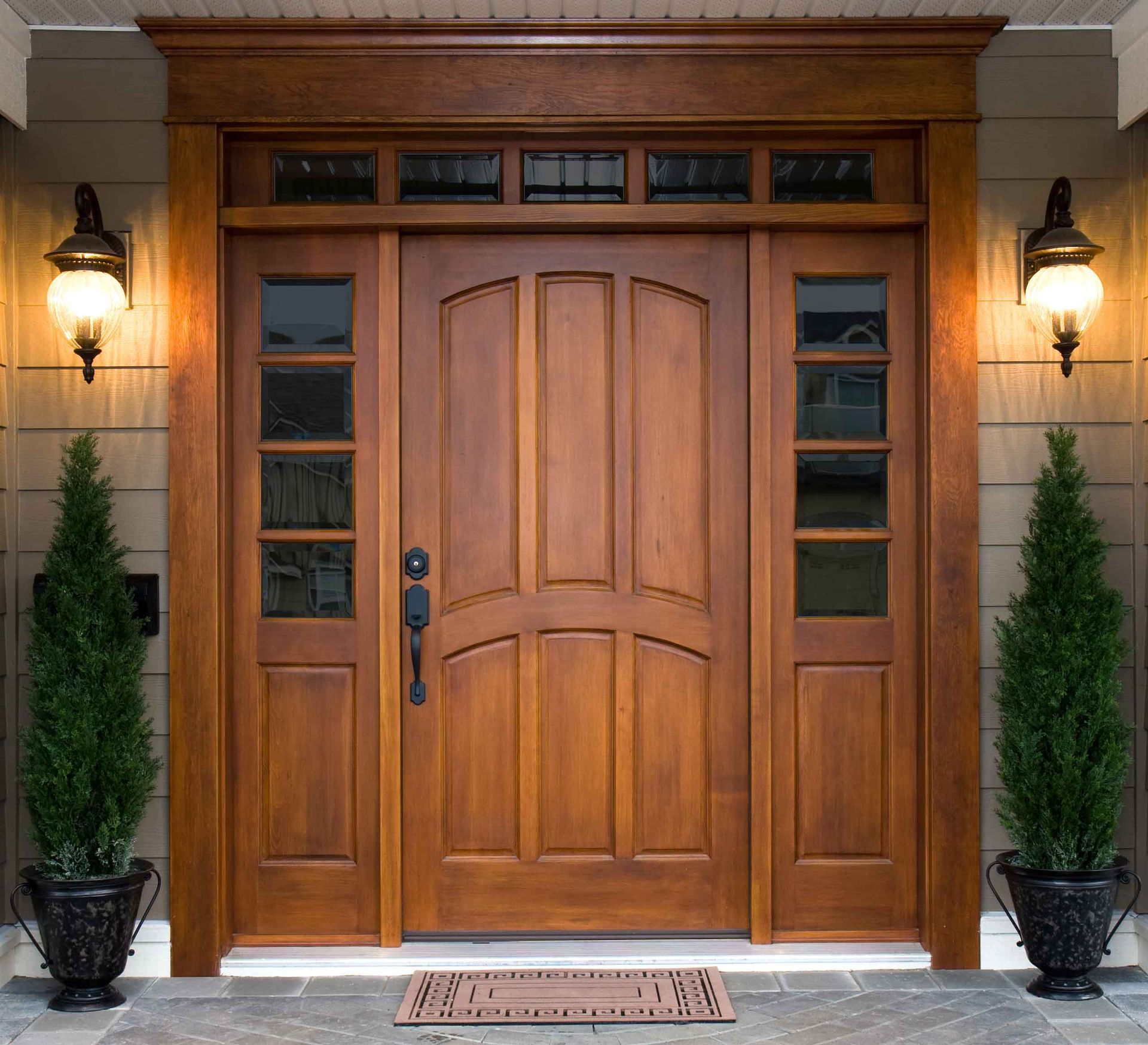 Wooden front door with sidelights and transom, flanked by lanterns and potted evergreens.